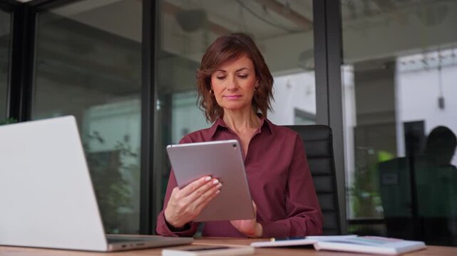 Businesswoman in 50s using digital tablet at office desk with copy space. Mature woman economist browses web on device, checks market email. Female business analyst reads reviews at workplace.