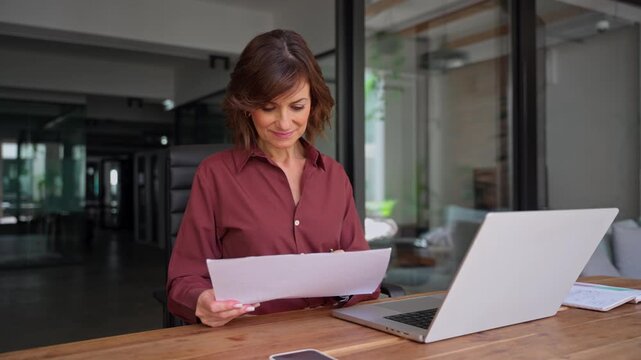 Latin businesswoman checking paper report at desk in office with copy space. Mature woman financial analyst holds document, works on laptop. Female expert reviews business contract at workplace.