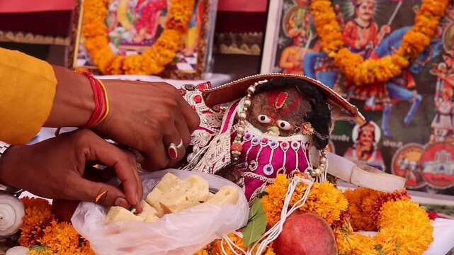 Close-up of a devotee's hands offering traditional Indian sweets as prasad to a beautifully decorated Hindu deity during a religious puja ceremony, surrounded by marigold flowers.
