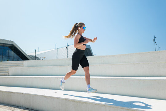 Running with energy under the bright sun, a woman showcases her fitness routine on a modern staircase