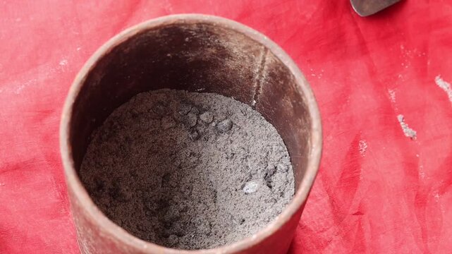 .
Close-up high angle shot of an Indian woman wearing glass bangles using a rustic metal mortar and pestle to traditionally crush and grind black salt against a red background.
