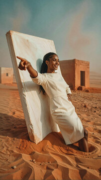 Woman in cream dress leans against monolithic stone slab while pointing toward horizon under warm sunlight within desert landscape with sandstone structure nearby.