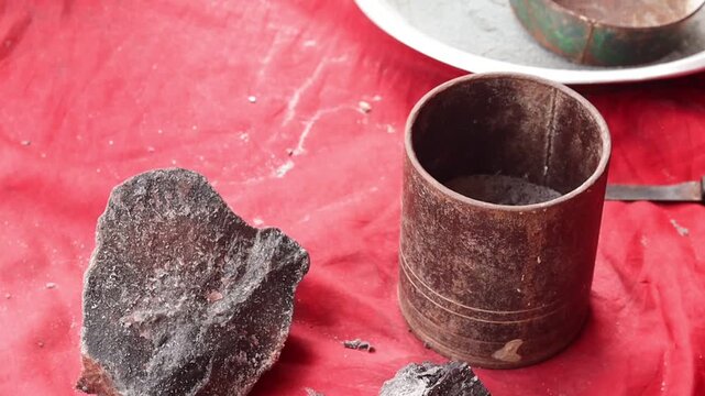 High angle shot of an Indian woman wearing green bangles using a rustic metal mortar and pestle to traditionally grind raw chunks of black salt against a red background.
