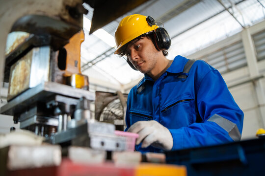 Skilled blue collar machine operator wearing yellow helmet and ear muffs carefully monitoring heavy metal press machine operation in modern industrial manufacturing plant facility.