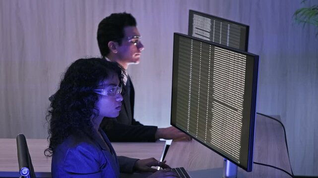 Two professionals code at large monitors filled with text in a blue-lit office, focused on software and security work.