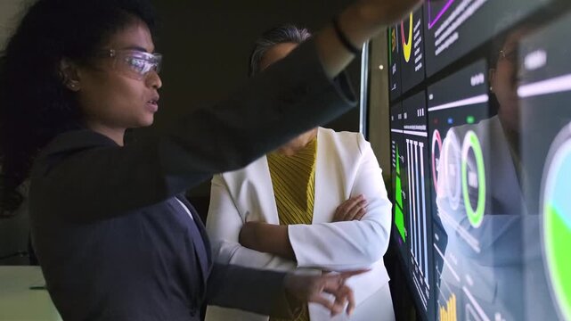Two professionals collaborate at a large interactive touchscreen, analyzing data dashboards and charts in a modern office and discussing insights.