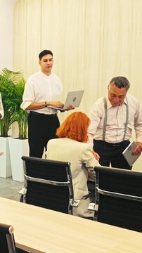 Colleagues gather in a meeting room as one person signs a document, while others wait with a tablet and notebook beside modern conference chairs and a light curtain backdrop.