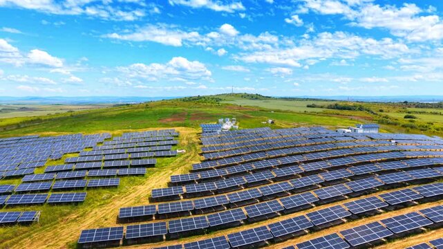 Parallel lines of photovoltaic arrays on a green field. Side aerial view of solar panels on a grassy meadow with dirt tracks under bright blue sky.