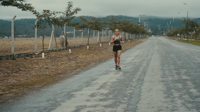 Wide shot of woman jogging on empty rural road past distant hills