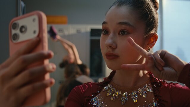 Young Female Uses Smartphone Front Camera as Mirror, Checking Makeup Before Training or Performance. Girl Smiles and Makes Peace Sign For Selfie in Locker Room. Youth Lifestyle, Social Media Culture.