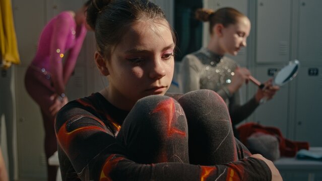Young Athlete in Crowded Dressing Room, Holding Knees Tightly With Sad, Anxious Expression, While Teammates Busy Preparing For Tournament. Intense Psychological Pressure, Stage Fright in Youth Sports.