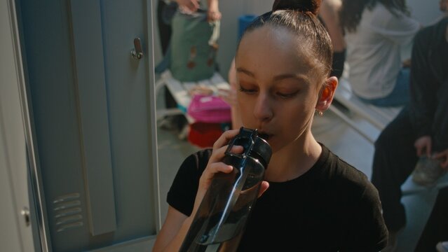 Focused Little Girl Drinking Water Next to Open Lockers in Dressing Room. She Does Light Warm Up Mobility Exercises, Preparing Body For Performance or Stretching Class. Healthy Lifestyle, Self Care.