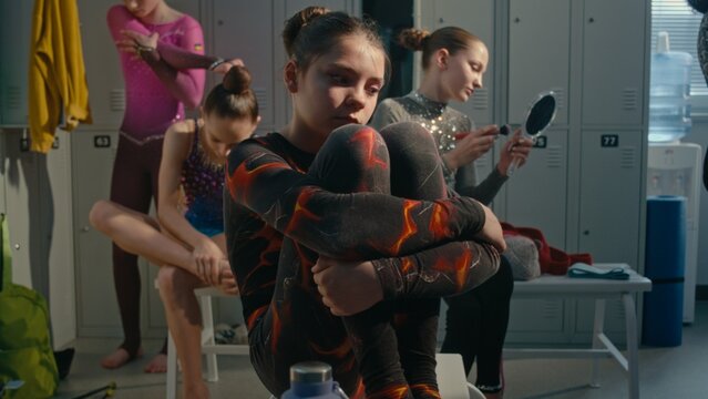 Young Athlete in Crowded Dressing Room, Holding Knees Tightly With Sad, Anxious Expression, While Teammates Busy Preparing For Tournament. Intense Psychological Pressure, Stage Fright in Youth Sports.