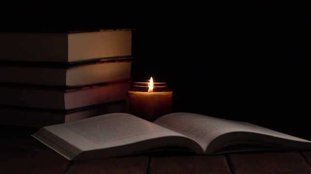 An open book lying on a wooden table with a burning candle beside it. Cozy atmosphere, reading, literature, study, and relaxation in warm candlelight