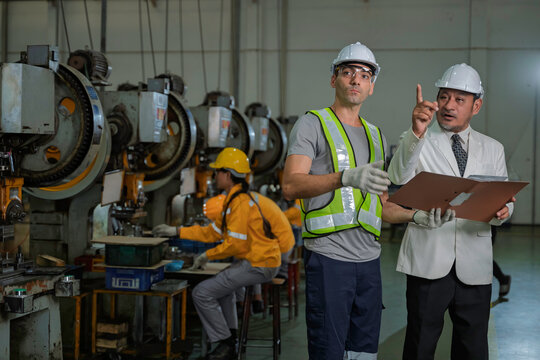 Senior Asian factory manager in a suit reviewing production documents with a male industrial engineer wearing safety gear on the manufacturing floor with other workers in the background.