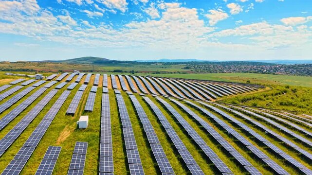 Aerial view of curved solar panel rows on hills. High angle drone shot of parallel curved rows of photovoltaic panels on a green field with a distant village.