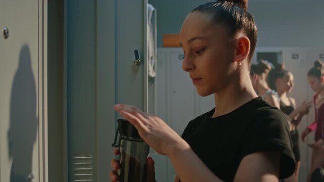 Young Athlete in Black Gymnastics Outfit Stands by Locker, Taking Refreshing Break to Drink Water From Sports Bottle. Girl Gently Stretches Neck, Shoulders, Recovering Energy Before Training Session.