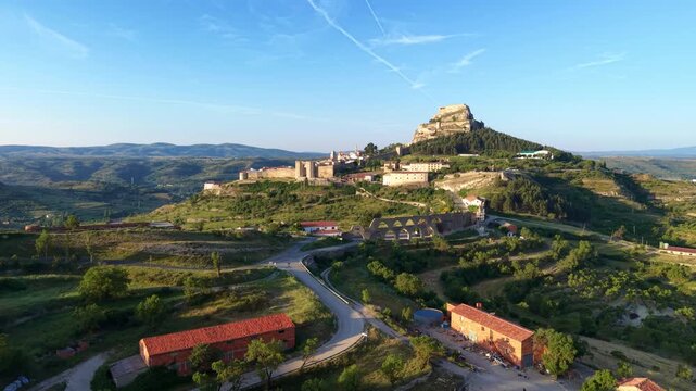 Aerial view of Morella's medieval castle crowning a rocky hilltop with fortified walls and the historic Els Arcs aqueduct arches visible, surrounded by green valleys in Castellon, Spain