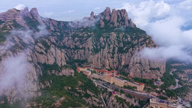 Aerial view of Santa Maria de Montserrat Abbey nestled among distinctive serrated rock formations with low clouds drifting through the Catalan mountain landscape