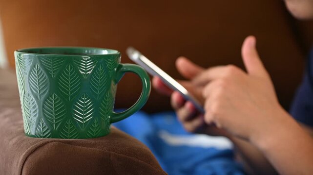 Green mug resting on sofa arm while person uses smartphone in living room