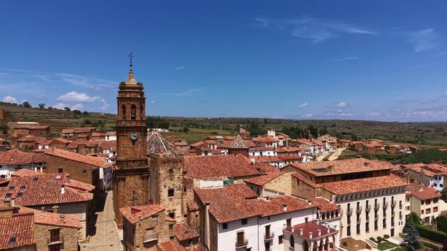 Elevated view of a baroque church bell tower with clock and cross rising above terracotta tile rooftops of La Iglesuela del Cid village, rolling countryside, Aragon, Spain