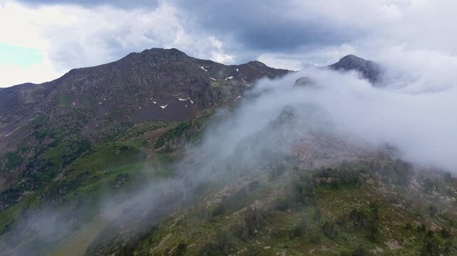 Low clouds cascade over a rugged rocky mountain ridge with patches of snow and green slopes in the high Pyrenees of Andorra, dramatic overcast sky above