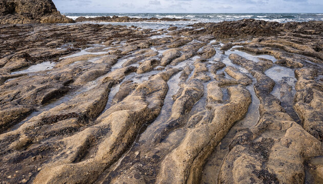 A rocky coastal shoreline with unique parallel grooves and ridges carved into the stone by water, leading towards the turbulent ocean waves