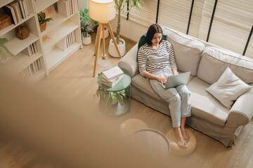 Overhead view of woman working remotely on laptop while relaxing on sofa, cozy living room with books, plants and natural light, casual work from home lifestyle scene with copy space © whyframeshot