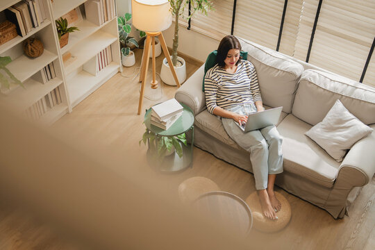 Overhead view of woman working remotely on laptop while relaxing on sofa, cozy living room with books, plants and natural light, casual work from home lifestyle scene with copy space