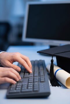 Graduate typing on keyboard with diploma and graduation cap on desk