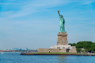 Statue of Liberty above the blue ocean in New York. Iconic green monument on its stone base with distant cargo ships and cranes on the clear horizon. © Vadim