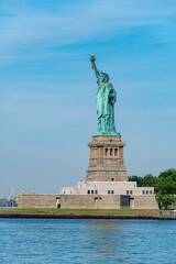 Statue of Liberty standing on Liberty Island in New York. Iconic copper monument stands tall on a stone pedestal surrounded by water under a bright clear sky. © Vadim