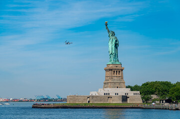 Statue of Liberty under blue sky with flying military airplane.. Profile of the monument with a tiltrotor aircraft in the sky. Calm ocean water fills the foreground. © Vadim