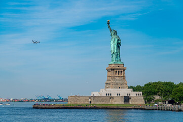 Statue of Liberty in New York with military aircraft flying.. Wide angle shot of the green copper monument under blue sky. A tiltrotor plane flies over the harbor water. © Vadim
