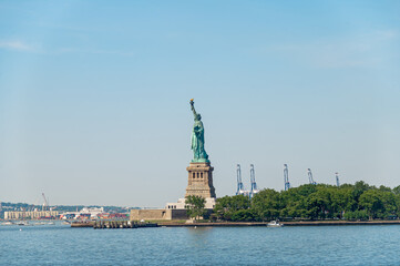 Statue of Liberty viewed from a boat in New York.. Long distance view of the iconic landmark on its base. Green trees and port cranes line the horizon. © Vadim