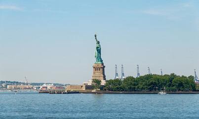 Statue of Liberty standing on Liberty Island in New York.. Full figure of the green copper sculpture from the water. Clear blue sky and industrial cranes behind. © Vadim