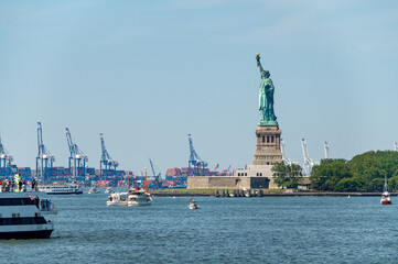Statue of Liberty overlooks harbor traffic on Independence Day. Ferries and small boats cross blue water with New Jersey container cranes behind Liberty Island. © Vadim