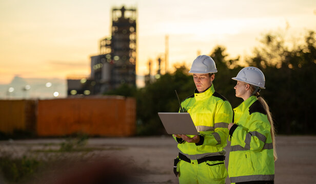 Engineers collaborating on construction site at sunset with safety gear and laptop in industrial environment
