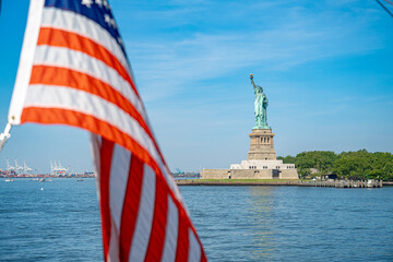 Statue of Liberty framed by American flag in harbor. A blurred striped banner fills the foreground beside Liberty Island under a clear blue sky. © Vadim