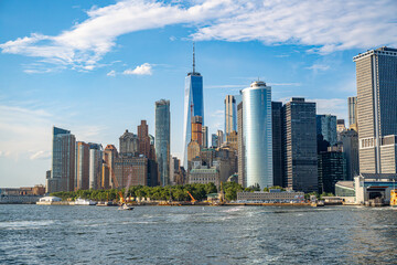 Obraz premium Lower Manhattan skyline and One World Trade Center waterfront. Office towers rise above trees and piers beside New York Harbor under scattered clouds.