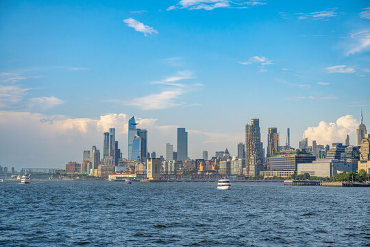 Midtown Manhattan skyline with Hudson Yards beside Hudson River. Ferries cross the water below the Empire State Building and waterfront towers under scattered clouds.