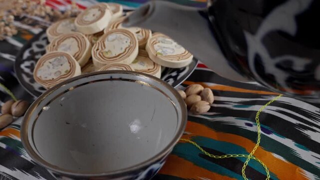 A hand pours tea from a traditional Uzbek teapot into a small teacup on colorful atlas fabric, with a plate of halva nearby
