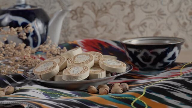 Traditional Uzbek halva topped with pistachios, served on a hand-painted ceramic plate alongside a steaming teapot and tea bowl, all arranged on vibrant Uzbek textile