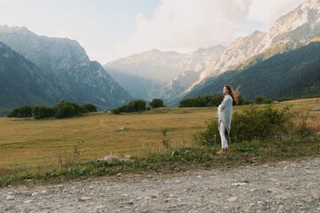 Obraz premium A woman in casual clothes stands on a dirt path in a vast mountain valley surrounded by green trees and rocky peaks under a cloudy sky. Lifestyle concept outdoors.