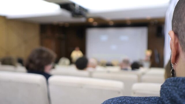 Audience watches presentation from back row. Focus on back of viewer's head. Blurred projection screen shows whiteboard. Attendees sit upright, attentive. Ideal for corporate events, training sessions