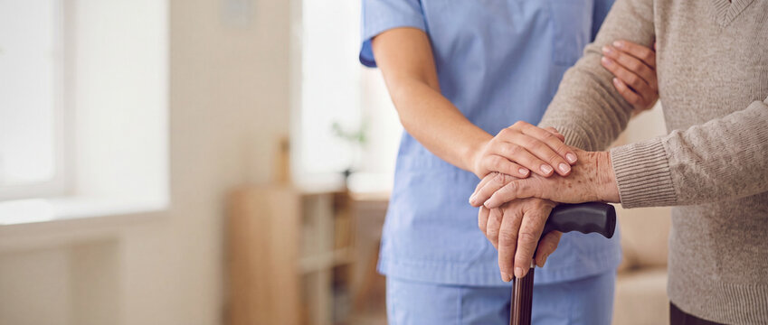 Professional medical caregiver supports elderly person hands resting on walking cane, copy space