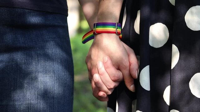 Close-up of two women holding hands. Rainbow bracelets (LGBT symbols) on the wrists of the girls. Concept of love, unity and support. Same-sex couple holding hands while walking