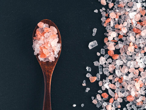Coarse pink himalayan salt crystals in a dark wooden spoon and scattered on black slate stone background. Top view flat lay composition with copy space. Perfect for cooking, spa or wellness concept