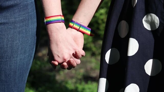 Same-sex couple holding hands while walking in the park. Focus on rainbow Pride bracelets as a symbol of equality. Concept of love, unity and support. LGBTQ+ couple holding hands