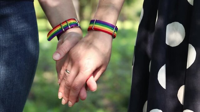 Close-up of two women holding hands. Rainbow bracelets (LGBT symbols) on the wrists of the girls. Concept of love, unity and support. Same-sex couple holding hands while walking
